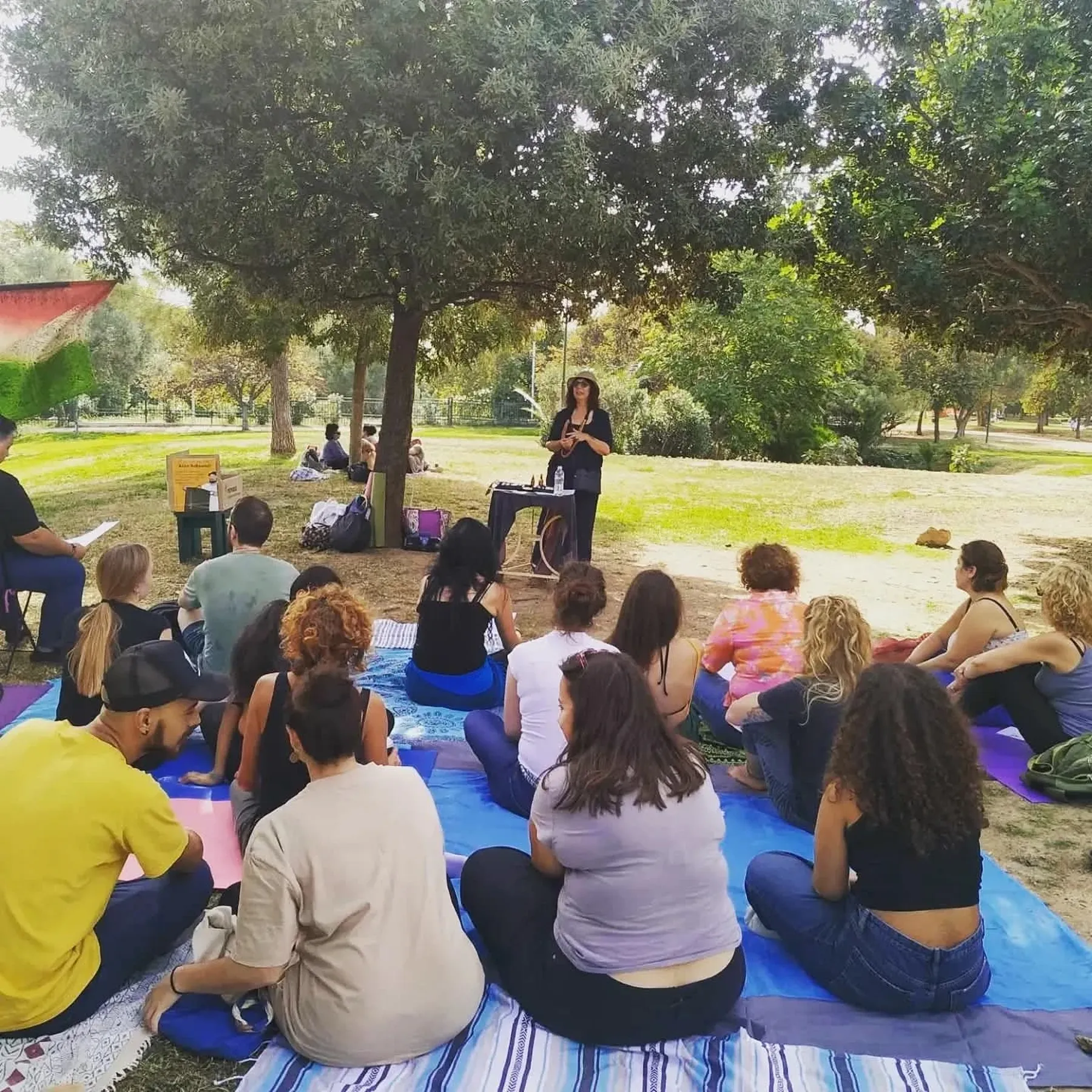 Συμμετέχοντες ακούν εισηγήτρια κάτω από τα δέντρα